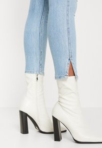 Botas blancas de tacón alto con una textura suave y brillante, con un cierre lateral y un tacón negro grueso. Llevadas con unos jeans ajustados de color azul claro.