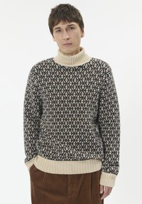 Mladić nosi džemper s uzorkom u mornarsko plavoj i bež boji te smešene pamučne hlače, stojeći protiv bijele pozadine.