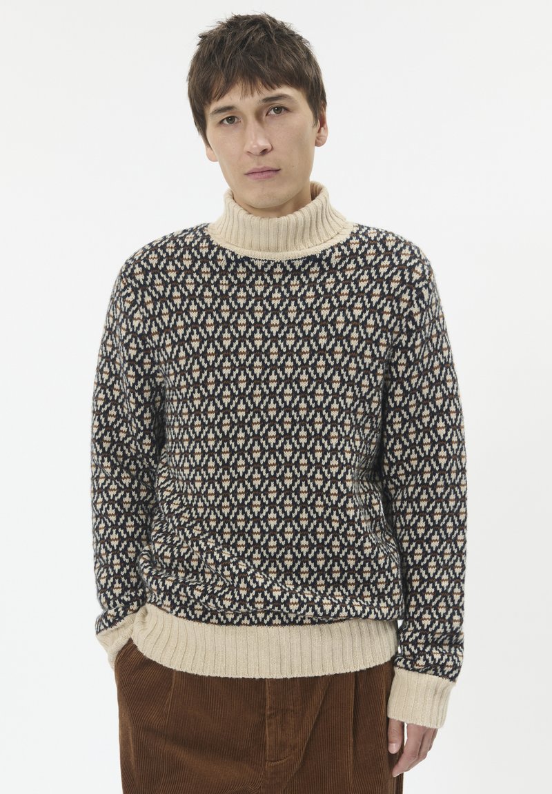 Mladić nosi džemper s uzorkom u mornarsko plavoj i bež boji te smešene pamučne hlače, stojeći protiv bijele pozadine.