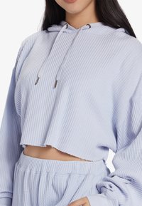 Hellblauera Cropped-Hoodie aus strukturiertem Stoff, mit Kordelzugkapuze, weiten Ärmeln und rohem Saum, kombiniert mit passenden strukturierten Hosen.