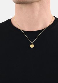 Collana con pendente a forma di cuore in oro, su una sottile catena, con dettagli incisi, indossata sopra una maglietta nera. Design semplice con una finitura lucida.