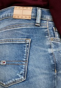 Gaiši zili džinsi ar nodilušu audumu. Tai ir ādas birka ar uzrakstu "TOMMY JEANS" un neliels logotipa akcents uz kabatas.