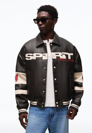 Man draagt een zwart varsityjack met witte strepen, tekst "SPIRIT", nummer 24 op de mouw, wit overhemd, blauwe jeans en zwarte zonnebril.