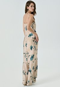 Robe longue beige avec des motifs floraux teal, fabriquée en tissu lisse et léger. Elle possède de fines bretelles et une silhouette fluide avec un ourlet évasé.