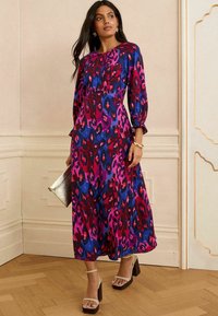 Farbenfrohes, langärmeliges Midi-Kleid im lila, rosa und blauen Leopardenmuster; mit rundem Ausschnitt und fließender Silhouette; kombiniert mit klobigen Absätzen.