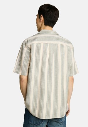 Mann mit dem Rücken zur Kamera trägt beige-graues, vertikal gestreiftes Kurzarmhemd und blaue Jeans vor schlichtem Hintergrund.