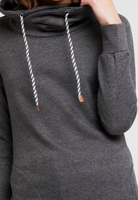 Szara bluza z kapturem o wysokim, złożonym kołnierzu, z białymi i czarnymi sznurkami oraz kieszenią kangurką. Miękki, teksturowany materiał.