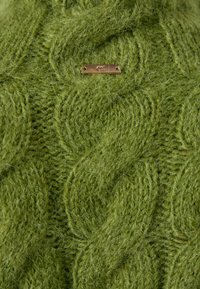 Pull vert en tricot avec un motif de câble texturé et une petite étiquette en métal. Matière en laine douce et duveteuse.