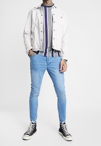 Chaqueta de denim blanca, camisa a rayas gris y morada, jeans ajustados de color azul claro y zapatillas altas negras. Estilo casual con texturas variadas.