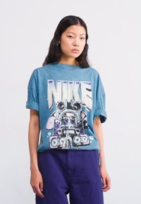 Per didelė mėlyna medvilninė marškinė su grafiniu dizainu, kuriame pavaizduoti garsiakalbiai ir Nike logotipas ryškiu baltu šriftu. Trumpomis rankovėmis ir laisva forma.