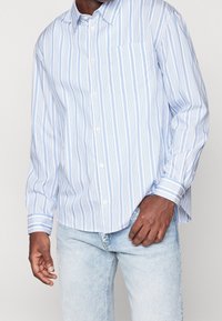 Světle modrá košile na knoflíky s vertikálními bílými pruhy, dlouhými rukávy, kapsou na prsou a zaobleným lemem; vyrobena z tkané látky.