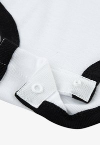 Indumento bianco con colletto e polsini neri a coste, caratterizzato da chiusura a scatto con bottoni rotondi bianchi e dettagli di cuciture a contrasto.