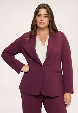 Donna con lunghi capelli mossi che indossa un blazer prugna aderente e pantaloni abbinati con una camicetta bianca, in piedi con una mano sui fianchi.