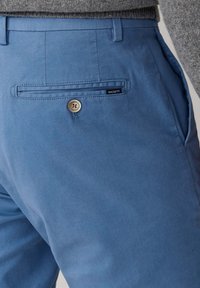 Lichtblauwe katoenen broek met een gladde textuur, voorzien van een achterzak met een knoop en een klein merkje.