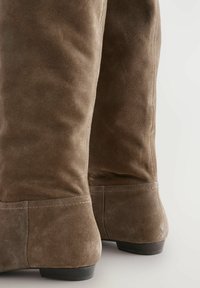 Botas marrones de ante hasta la rodilla con una textura suave, tacón cuadrado y detalles de costura sutiles a lo largo de las costuras.