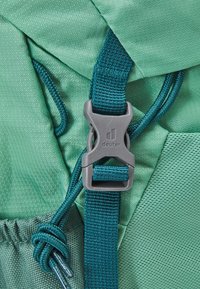 Mint green backpack fabric with a grey plastic buckle, accented by dark teal straps and cords. Textured surface detail visible.