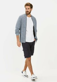 Blue textured zip-up jacket over a white t-shirt, paired with dark cargo shorts and white sneakers with navy accents.