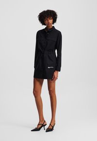 Abito camicia nero con maniche lunghe, colletto abbottonato, vita cinte, due tasche sul petto e dettaglio del logo sul davanti. Indossato con sandali neri con tacco.