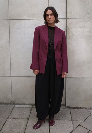 Burgundy tailored blazer over a black sheer top, paired with wide black trousers and burgundy heels. Smooth fabric, structured design.