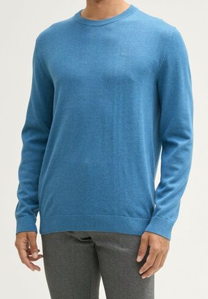 Homme portant un pull bleu à manches longues et col rond ainsi qu’un pantalon gris, debout devant un fond clair uni.