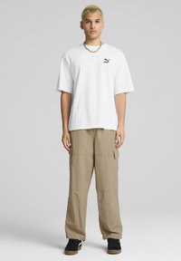 Witte t-shirt met korte mouwen en zwart Puma-logo, gecombineerd met beige cargobroek met zijzakken en zwarte schoenen met een gumrubberen zool.