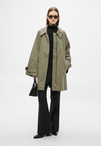 Manteau surdimensionné à motif pied-de-poule noir et crème, avec un large col, une fermeture à boutons et de grandes poches ; porté avec un pantalon noir et des lunettes de soleil.