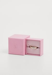 Rosafarbene Schmuckbox mit strukturiertem Oberflächen öffnet sich und enthüllt einen goldenen Ring mit fünf klaren Steinen in einer Reihe, der auf einem weichen Stoffkissen liegt.