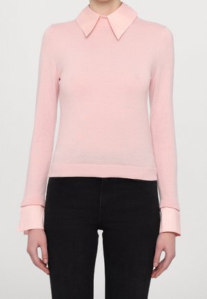Femme portant un pull rose clair à manches longues avec un col large et des poignets sur un pantalon noir, debout devant un fond uni.