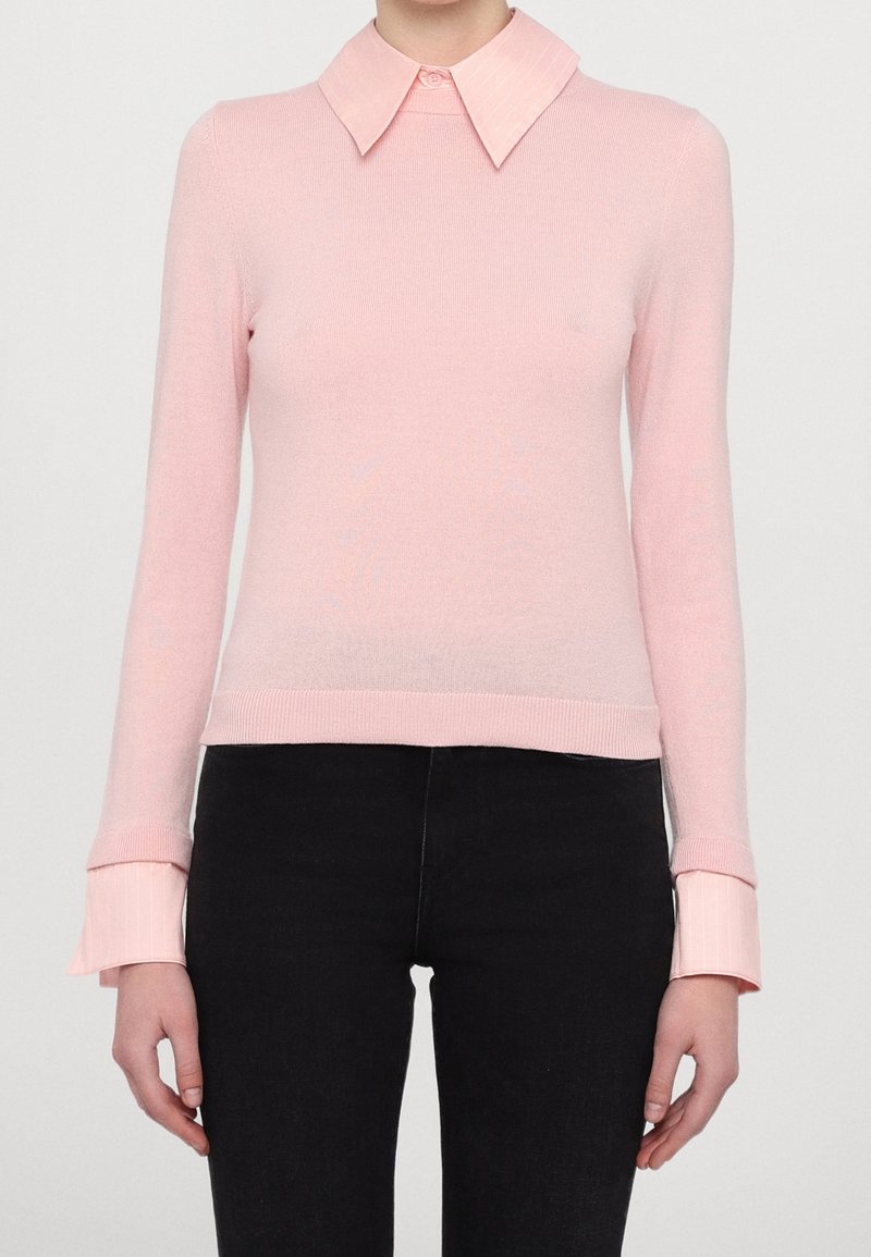 Femme portant un pull rose clair à manches longues avec un col large et des poignets sur un pantalon noir, debout devant un fond uni.