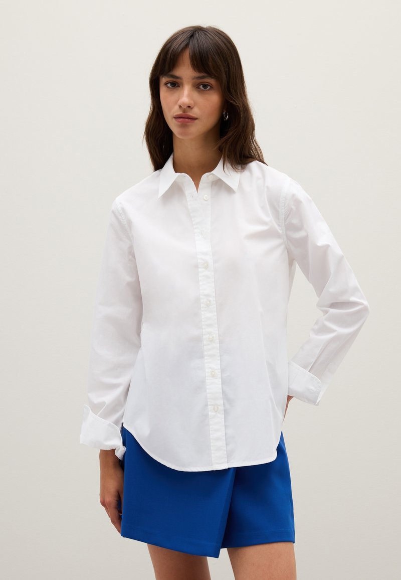 White button-up shirt made of cotton with long sleeves and a classic collar, paired with blue shorts. The shirt has a straight cut.