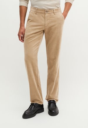 Mann trägt beige Cordhosen, schwarze Lederschuhe und ein hellbeiges Langarmhemd und steht mit den Händen in den Taschen.