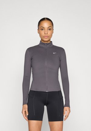 Veste de sport grise à manches longues avec un col montant, une fermeture éclair à l'avant et une coupe ajustée. Portée par-dessus un short noir avec poches latérales.