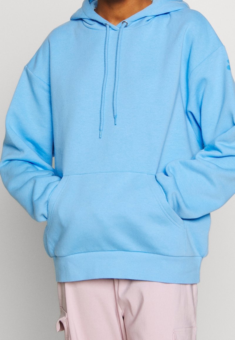 Personne portant un sweat à capuche bleu clair avec des cordons et une poche avant, associé à un pantalon cargo rose clair, debout devant un fond neutre.