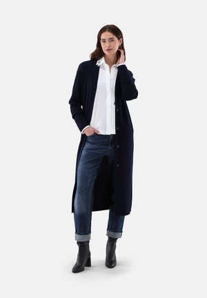 Navyfarbener gerippter langer Cardigan aus Strickstoff, mit Knopfverschluss, Seitenschlitzen, getragen über einem weißen Hemd mit Kragen und blauen Jeans.