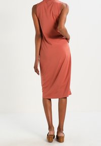 Kvinna klädd i ärmlös, korallfärgad midiklänning och bruna klackskor med blockklack, stående med ena handen på höften, ryggen mot kameran.