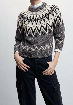 Persona con suéter de punto con patrón en negro, blanco y gris, y pantalones cargo negros, con las manos en las caderas y anillos dorados visibles.