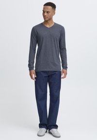 Grå langærmet V-hals skjorte, parret med løse blå jeans og grå sneakers, præsenteret på en stående mandlig model mod en hvid baggrund.
