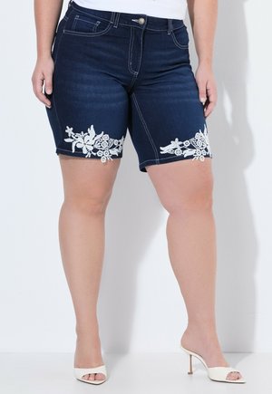 Donna che indossa shorts di denim blu scuro con dettagli in pizzo floreale bianco vicino all'orlo e sandali bianchi con tacco e punta aperta.