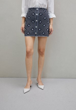 Wit shirt is in een high-waisted blauwe en zwarte mini-rok met patroon en knopen aan de voorkant gestopt, gecombineerd met witte pumps met puntige neus.