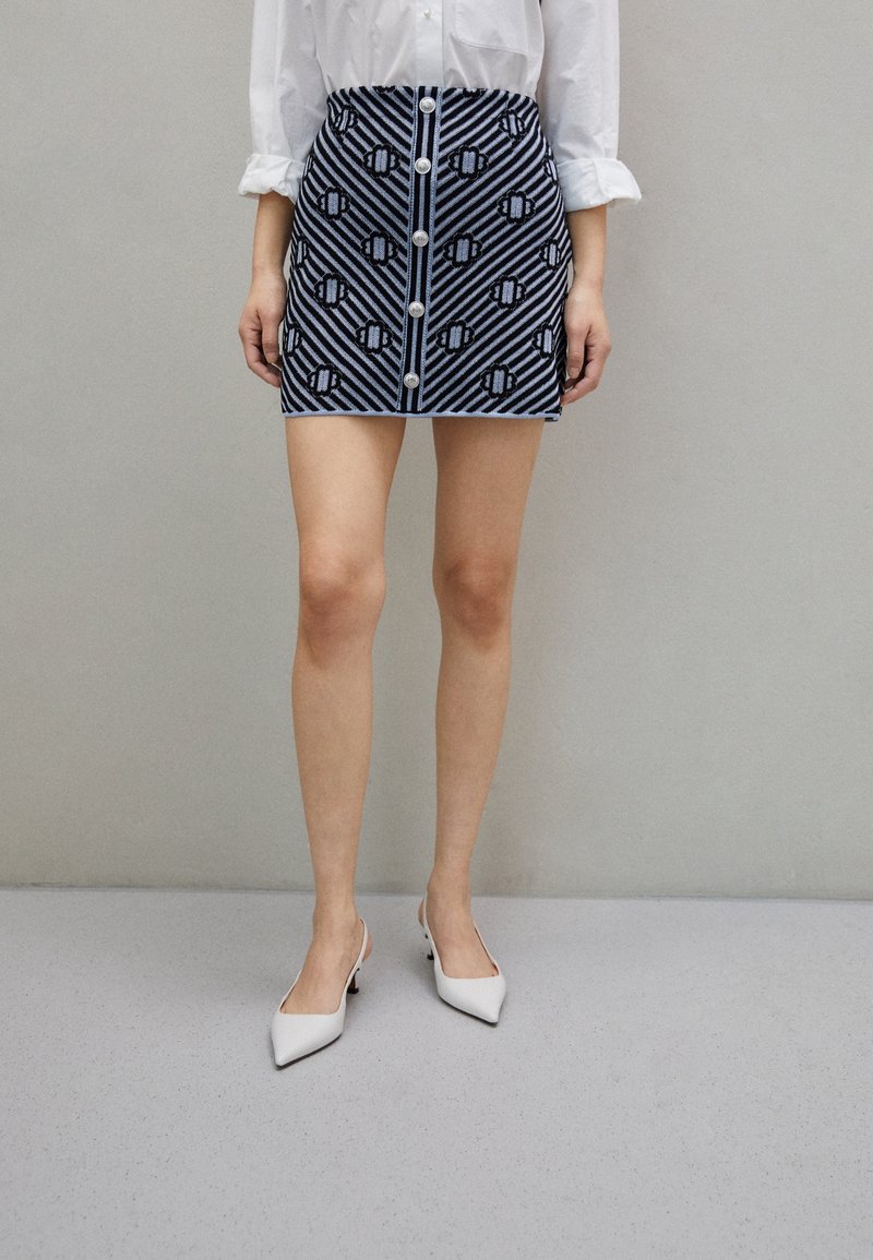 Une chemise blanche est rentrée dans une mini-jupe taille haute bleue et noire à motifs avec des boutons à l'avant, associée à des escarpins blancs à bouts pointus.