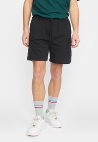 Homme portant un short cargo noir, des chaussettes grises avec des rayures rouges et bleues, et des baskets blanches, debout devant un fond uni.