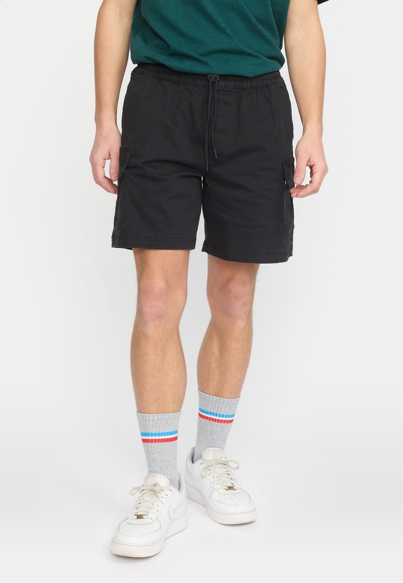 Homme portant un short cargo noir, des chaussettes grises avec des rayures rouges et bleues, et des baskets blanches, debout devant un fond uni.