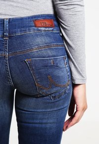 Smala blå denimjeans med orange sömmar, med en brun läderetikett och två bakfickor med dekorativ sömdetalj.