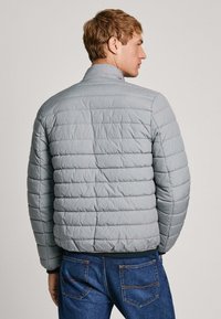 Sivá prešívaná puffa bunda s vysokým golierom, s horizontálnym prešívaním a rebrovaným lemom, v kombinácii s tmavomodrými džínami.