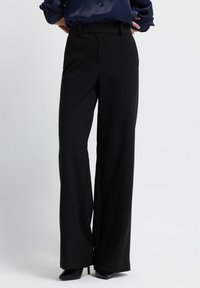 Femme portant un pantalon large taille haute noir et des escarpins noirs à bout pointu, se tenant les mains sur les hanches contre un fond neutre.