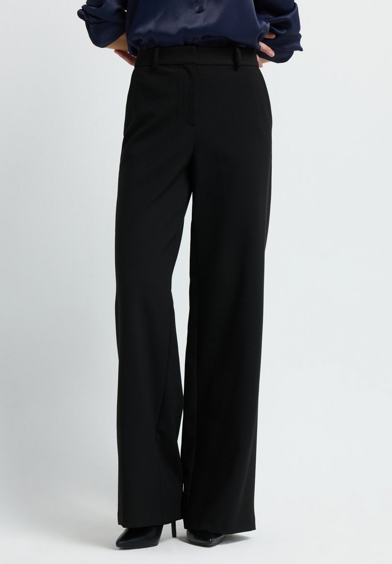 Femme portant un pantalon large taille haute noir et des escarpins noirs à bout pointu, se tenant les mains sur les hanches contre un fond neutre.
