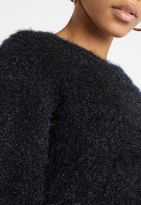 Black fuzzy knit sweater with a loose fit, featuring a textured surface and wide neckline. Close-up highlights soft fibers and rich color.