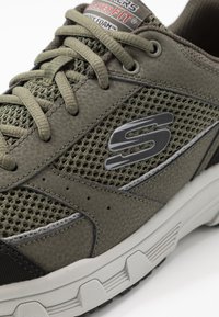 Zapatilla deportiva verde con paneles de malla, cuero texturizado y una suela de goma gruesa. Logo visible en el lado. Sistema de cordones elevado.