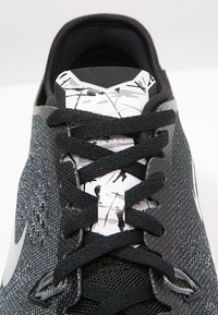 Sort atletiksko med en tekstureret mesh-overdel, der har sorte snørebånd, en stiliseret hvid tunge med sorte grafiske accenter og en reflekterende swoosh.
