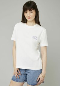 T-shirt en coton blanc à manches courtes avec un texte violet clair sur le côté gauche. Le tissu est lisse et légèrement structuré.